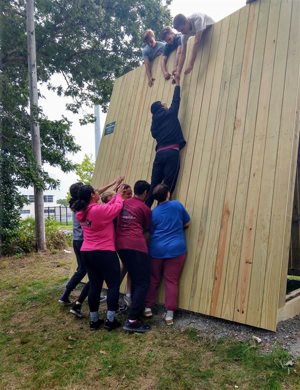 Students assisting a classmate up the free standing low wall in adventure education